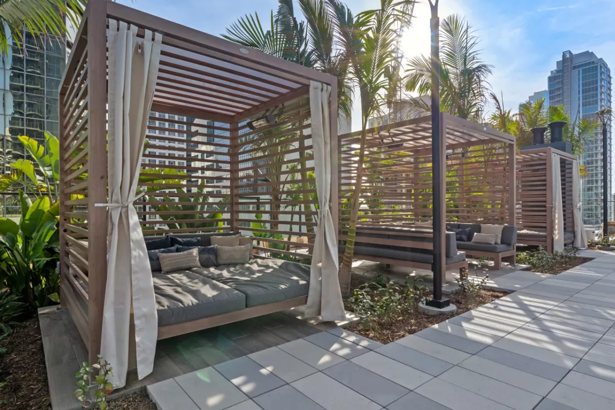 Wooden cabanas with cushioned seating, surrounded by palm trees and city buildings.