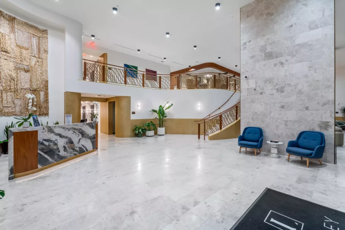 Modern lobby with marble floors, a curved staircase with golden railings, a textured wall art piece, and a marble-front desk.