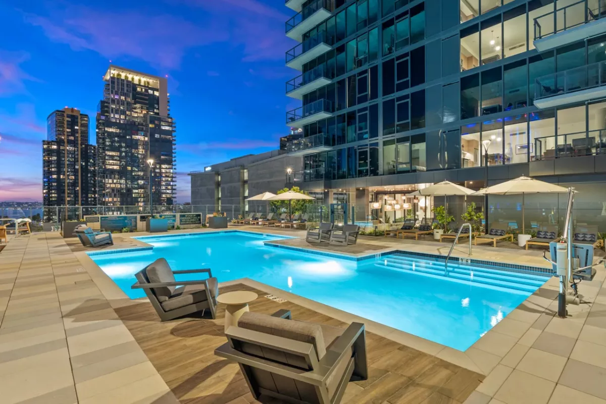  Illuminated rooftop pool at dusk with lounge chairs, umbrellas, and city skyline view.
