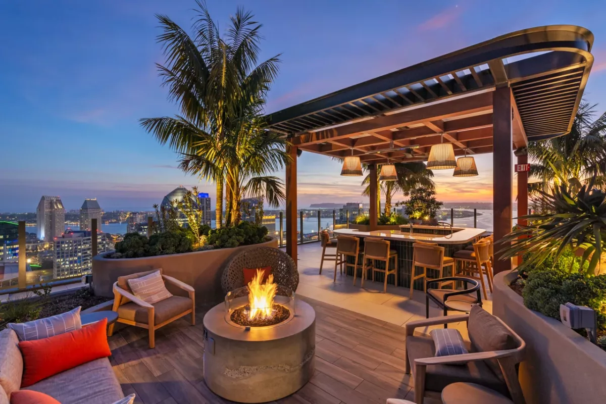  Rooftop lounge with fire pit, pergola bar, palm trees, and city skyline at sunset.