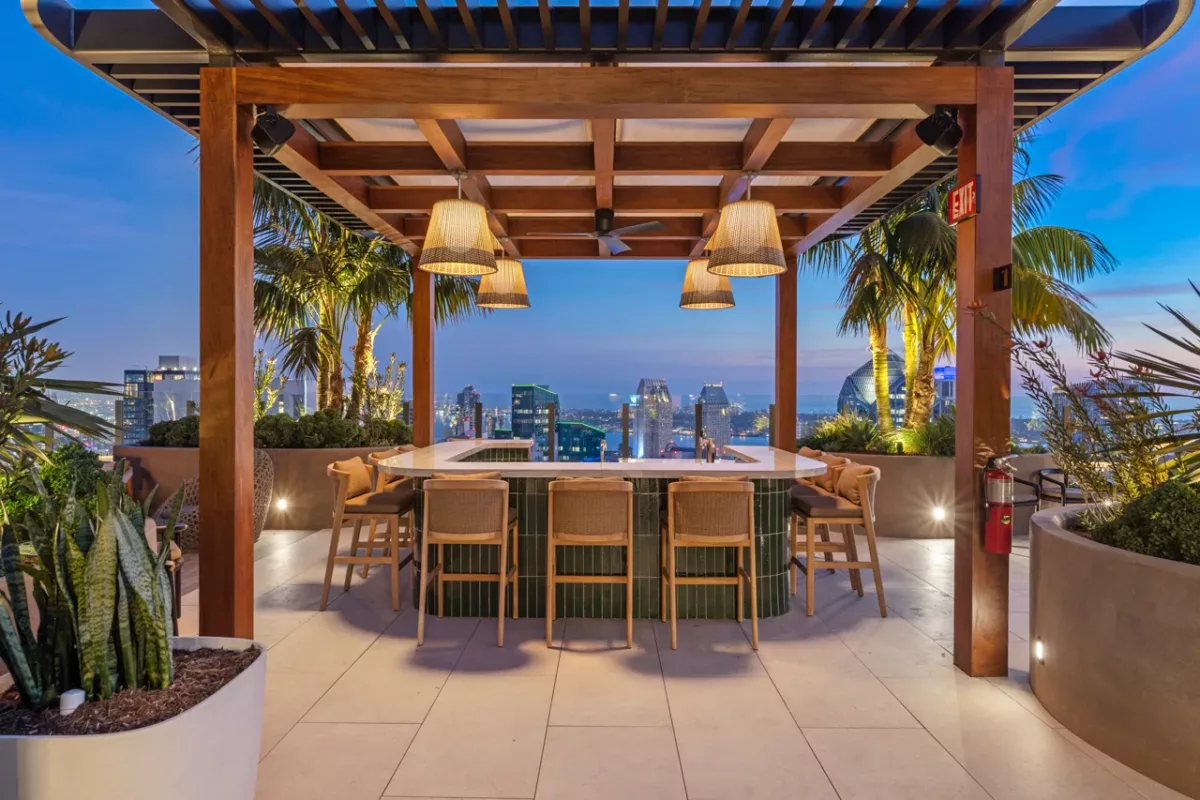 Rooftop bar with wood pergola, pendant lights, and skyline views framed by palm trees at dusk.