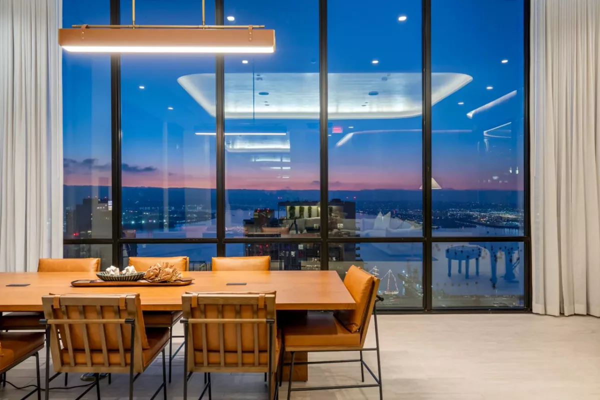 Dining table with leather chairs beside tall windows showing a dusk city and waterfront view.