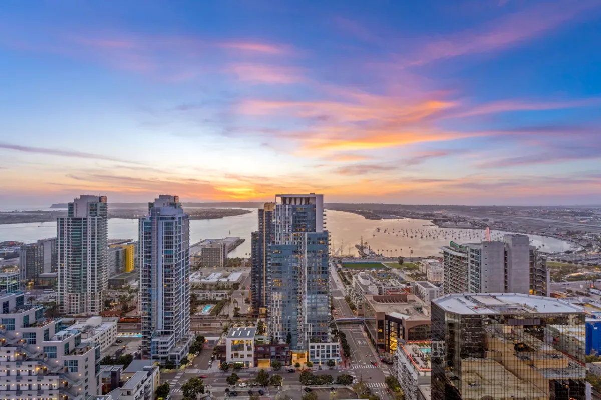 Panoramic cityscape at sunset with high-rises, harbor, and boats on calm water.