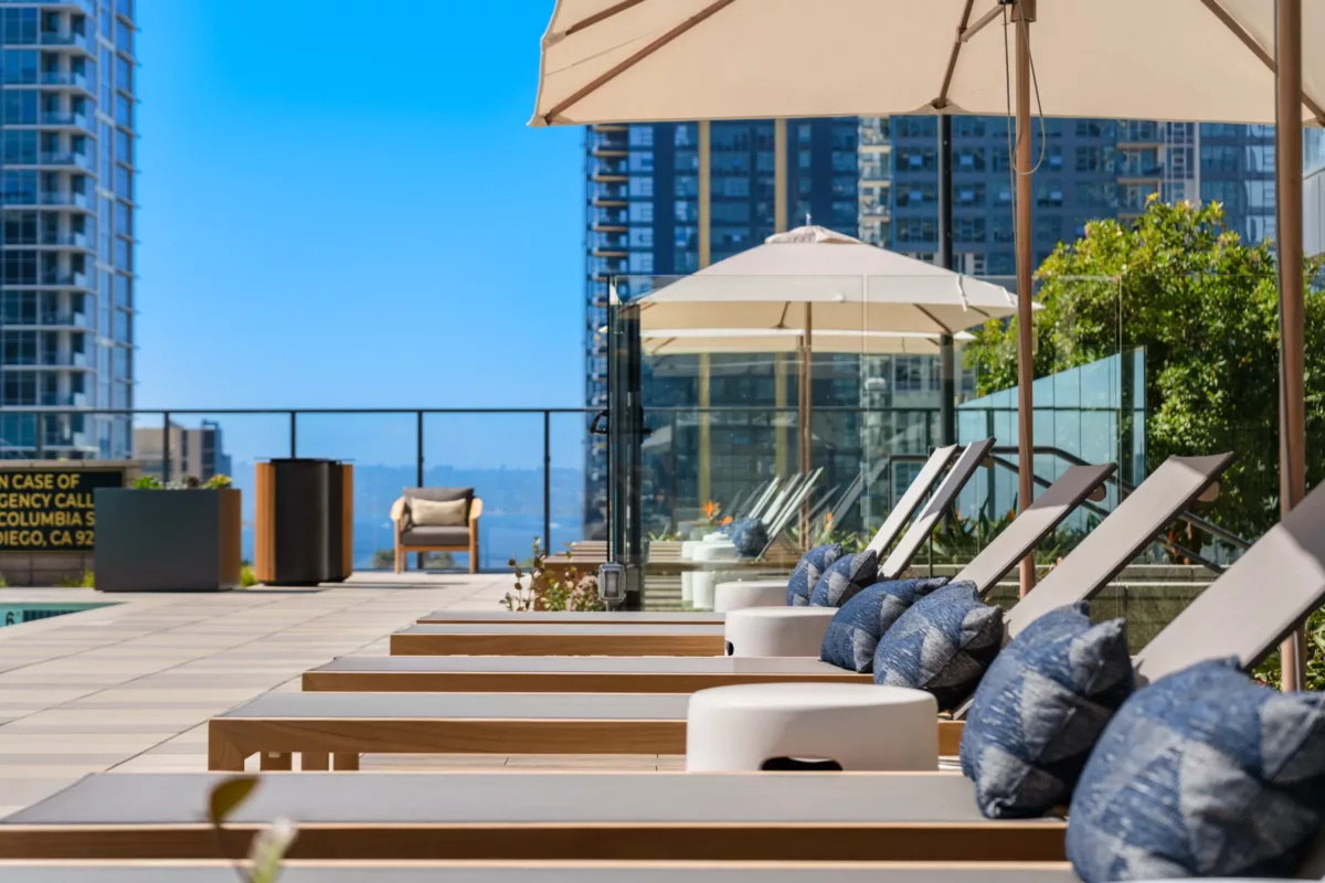 Row of poolside lounge chairs with umbrellas and pillows overlooking city skyline.