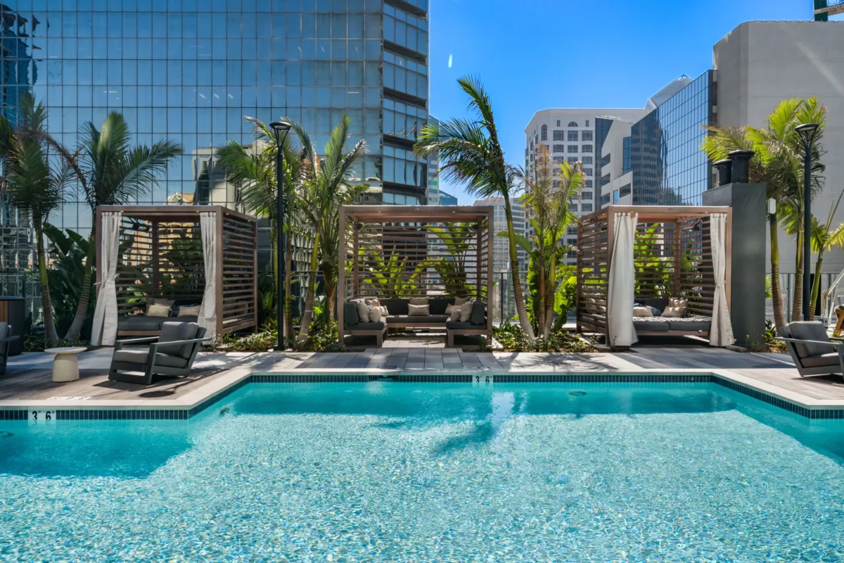 Outdoor pool with wooden cabanas, palm trees, and high-rise buildings in background.