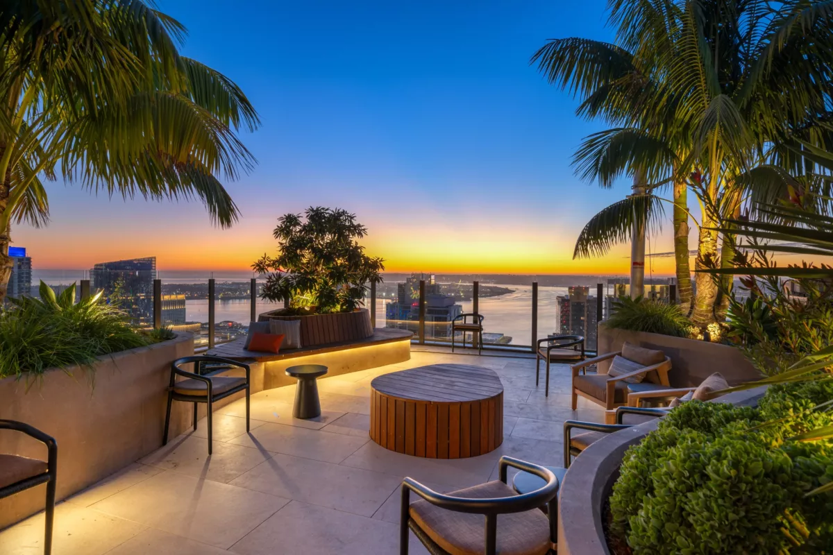 Rooftop terrace with modern seating and tropical plants overlooking bay and city at sunset.