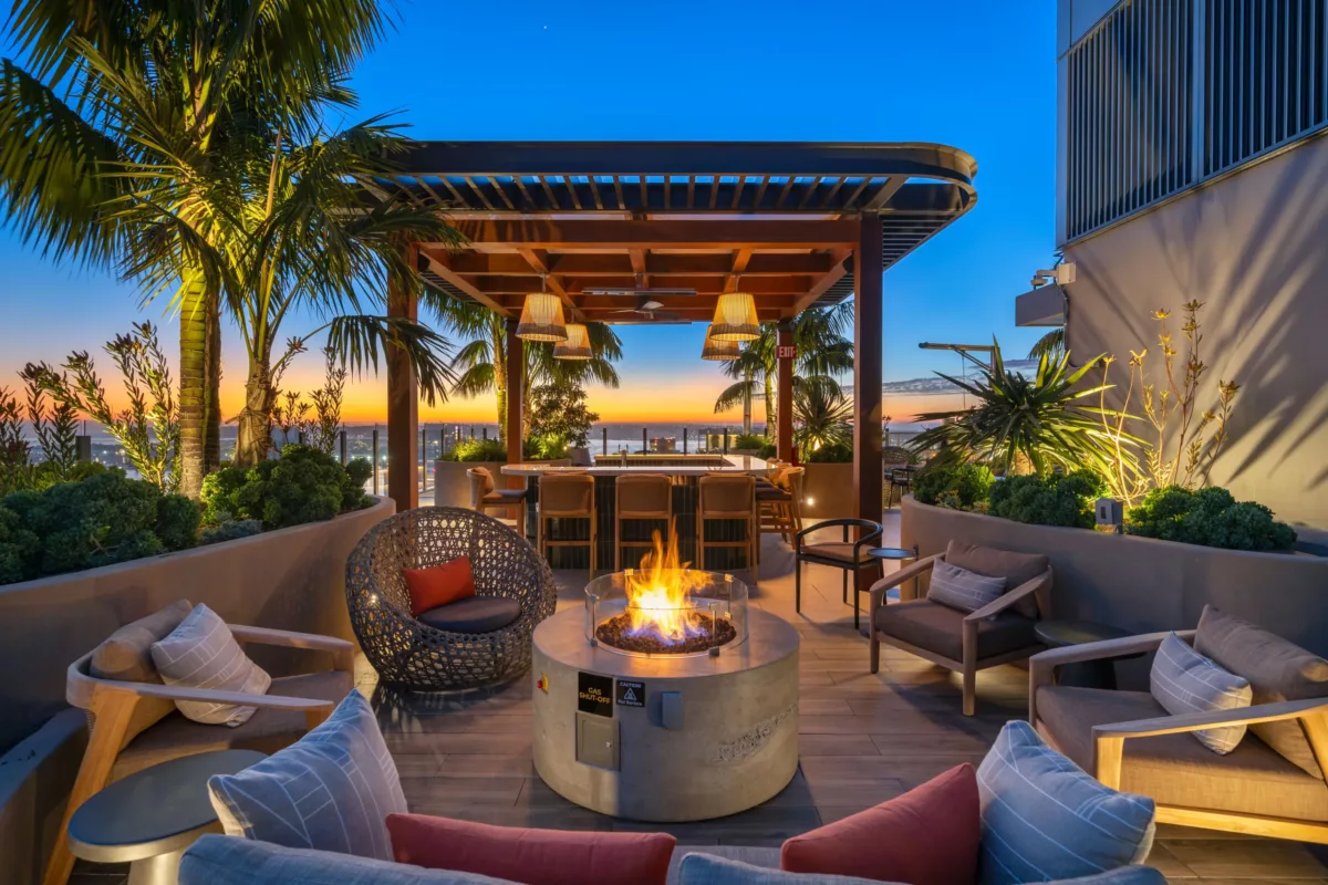 Cozy rooftop fire pit lounge with pergola bar, palm trees, and ocean sunset in the background.
