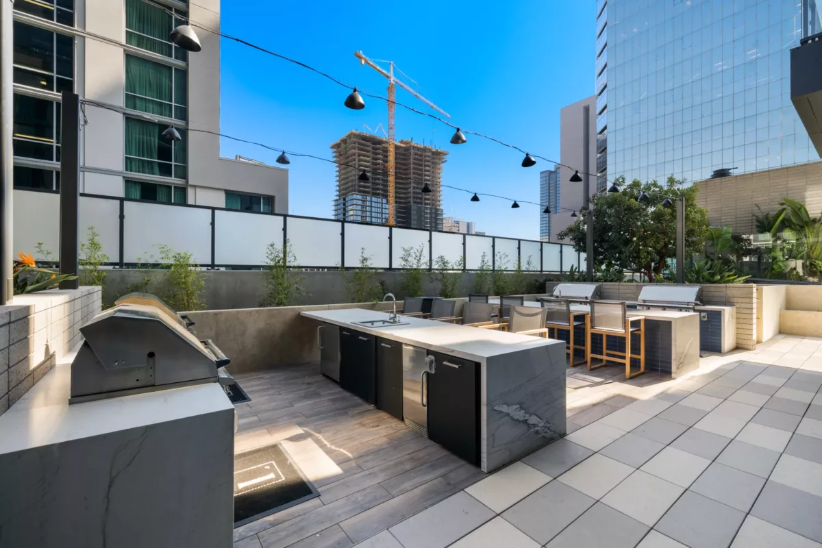 Urban patio with built-in grills, dining counters, and string lights under a clear blue sky.