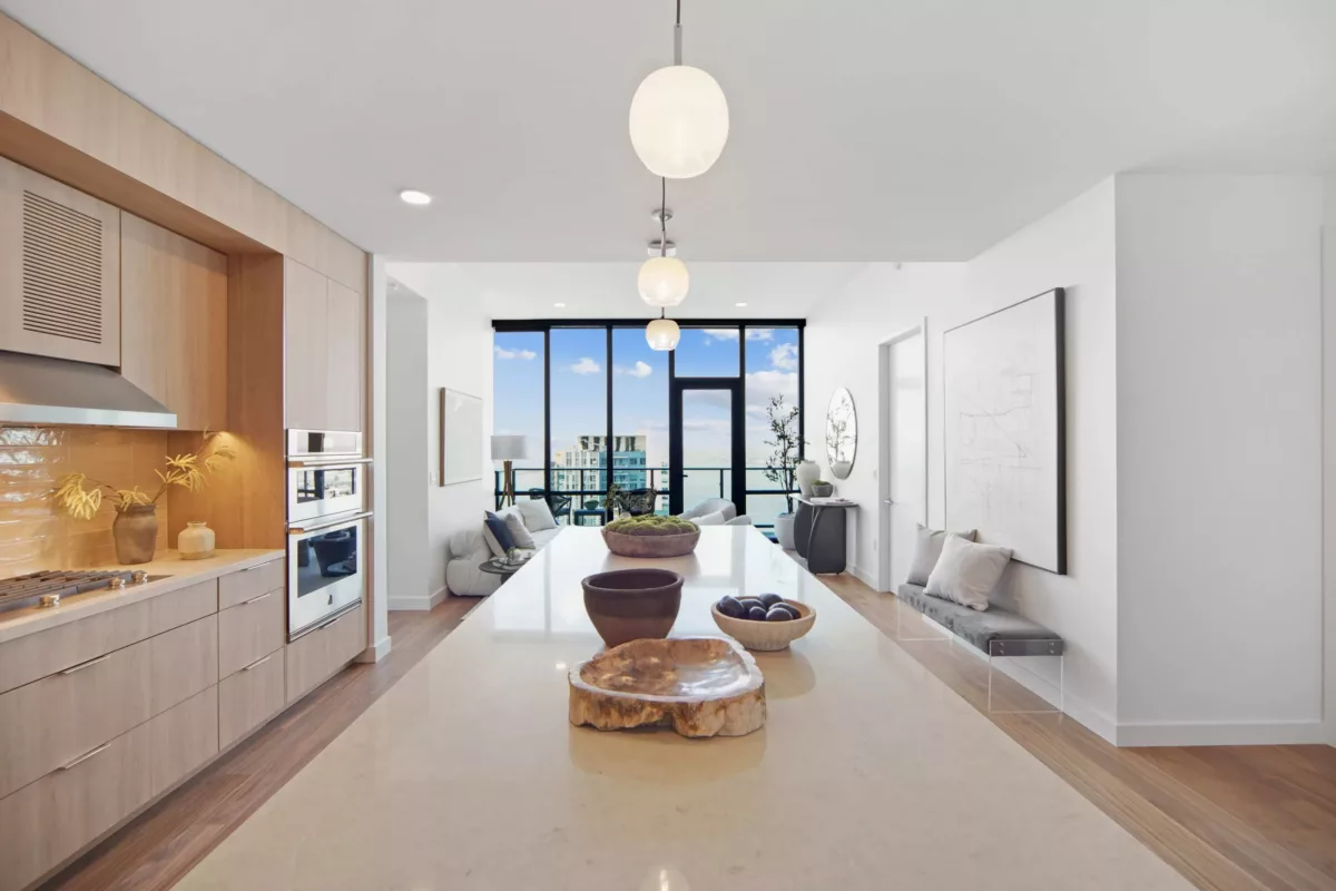 Open-concept kitchen with light wood-style cabinets, stainless steel appliances, and an island leading to a bright living area with large windows.