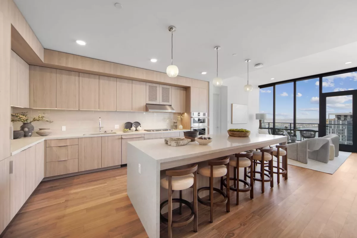Spacious kitchen with a central island, bar seating, and pendant lights, opening to a living area with large windows and city views.