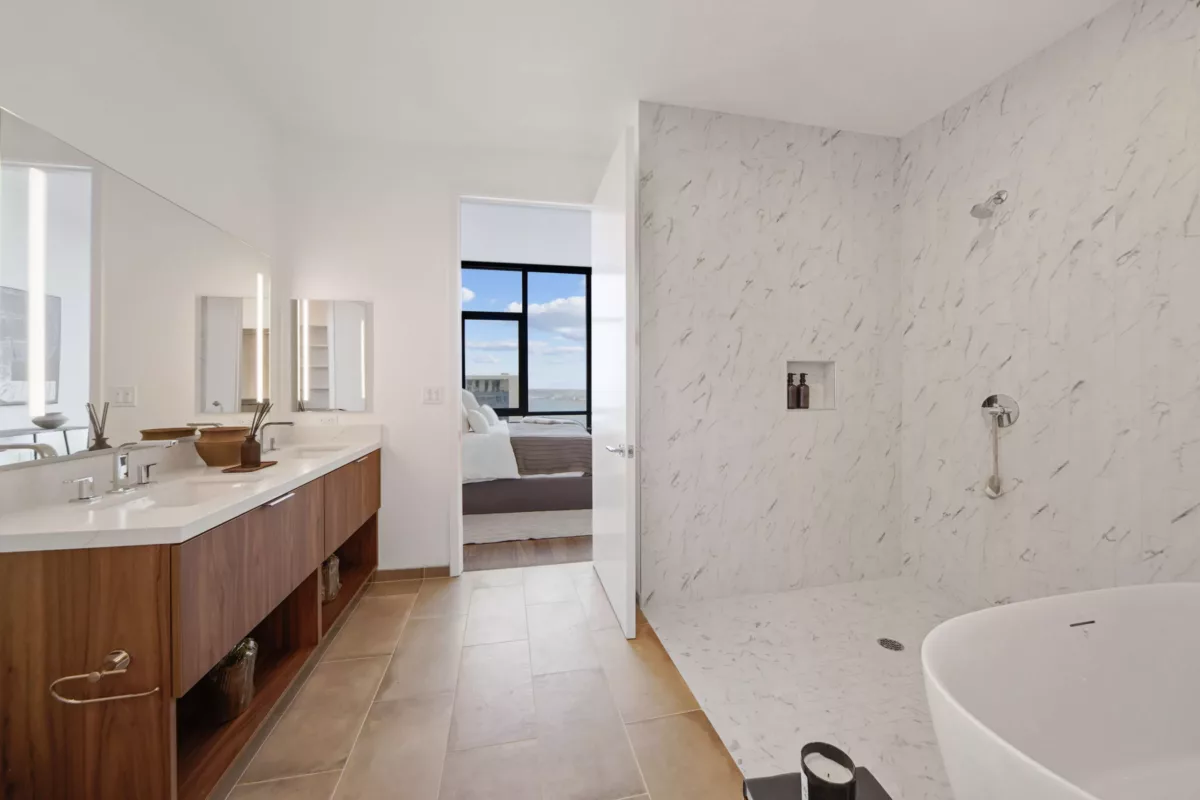 Modern bathroom with a double vanity, marble shower, soaking tub, and a view into the adjoining bedroom with large windows.
