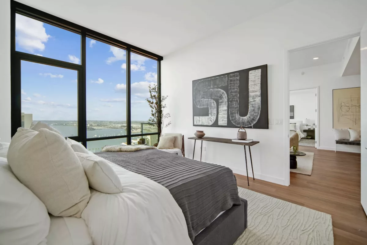 Modern bedroom with large windows offering a city and waterfront view, featuring a bed with a gray comforter, minimalist decor, and abstract wall art.