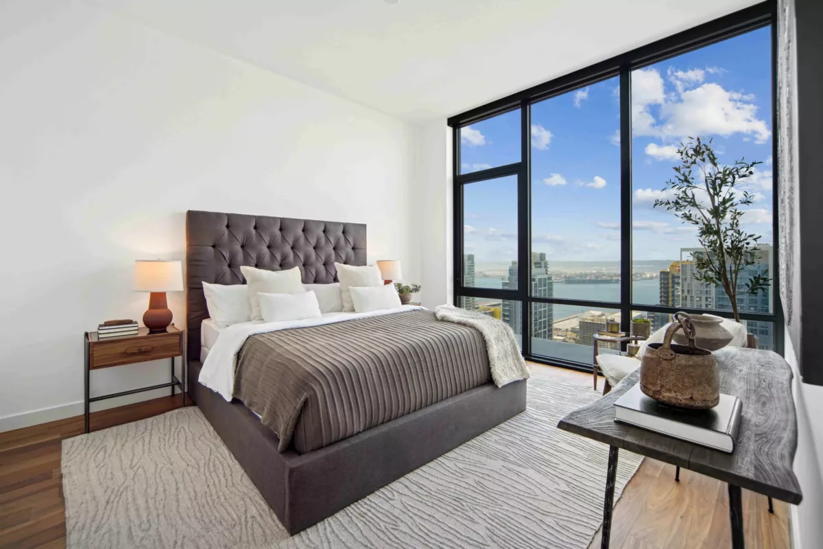 Stylish bedroom with a tufted headboard, layered bedding, and floor-to-ceiling windows showcasing a city skyline and waterfront.