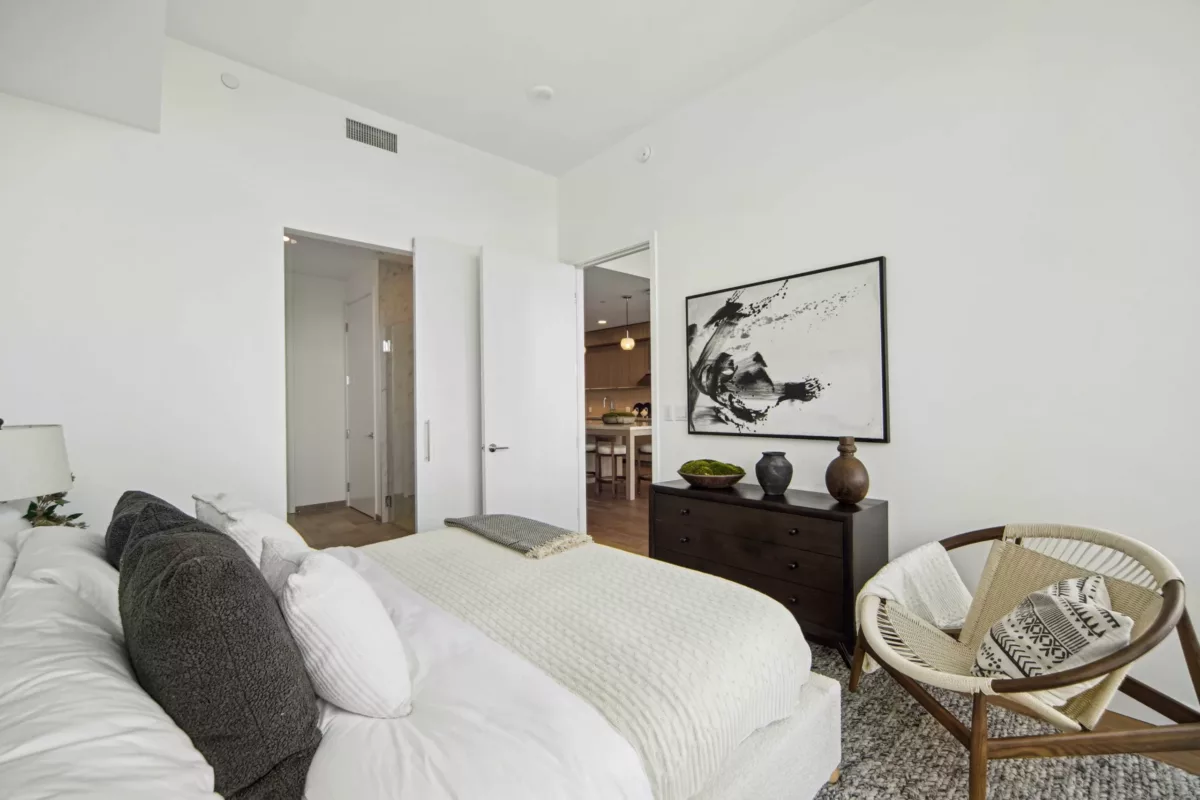 Cozy bedroom with a white bed, wicker chair, dark dresser, and abstract black-and-white artwork on a clean white wall.