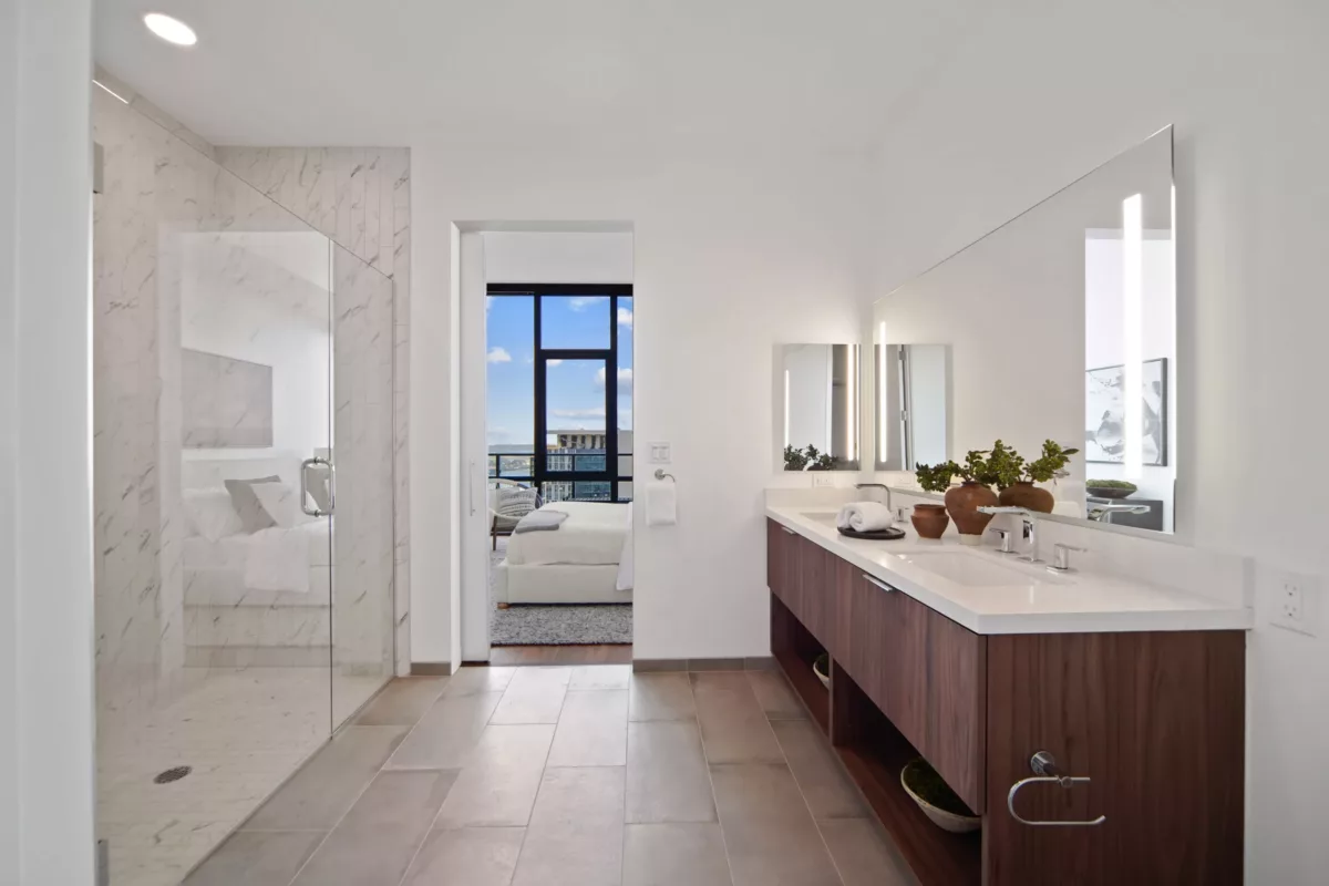 Bathroom with a marble-tiled shower, wood vanity, and a glimpse into the bedroom with large windows overlooking the city.