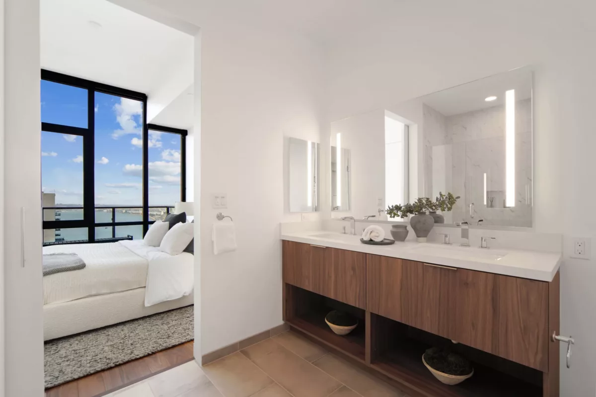Bright bathroom with a wood vanity, dual sinks, LED-lit mirror, and a view of a bedroom featuring large windows and a city skyline.