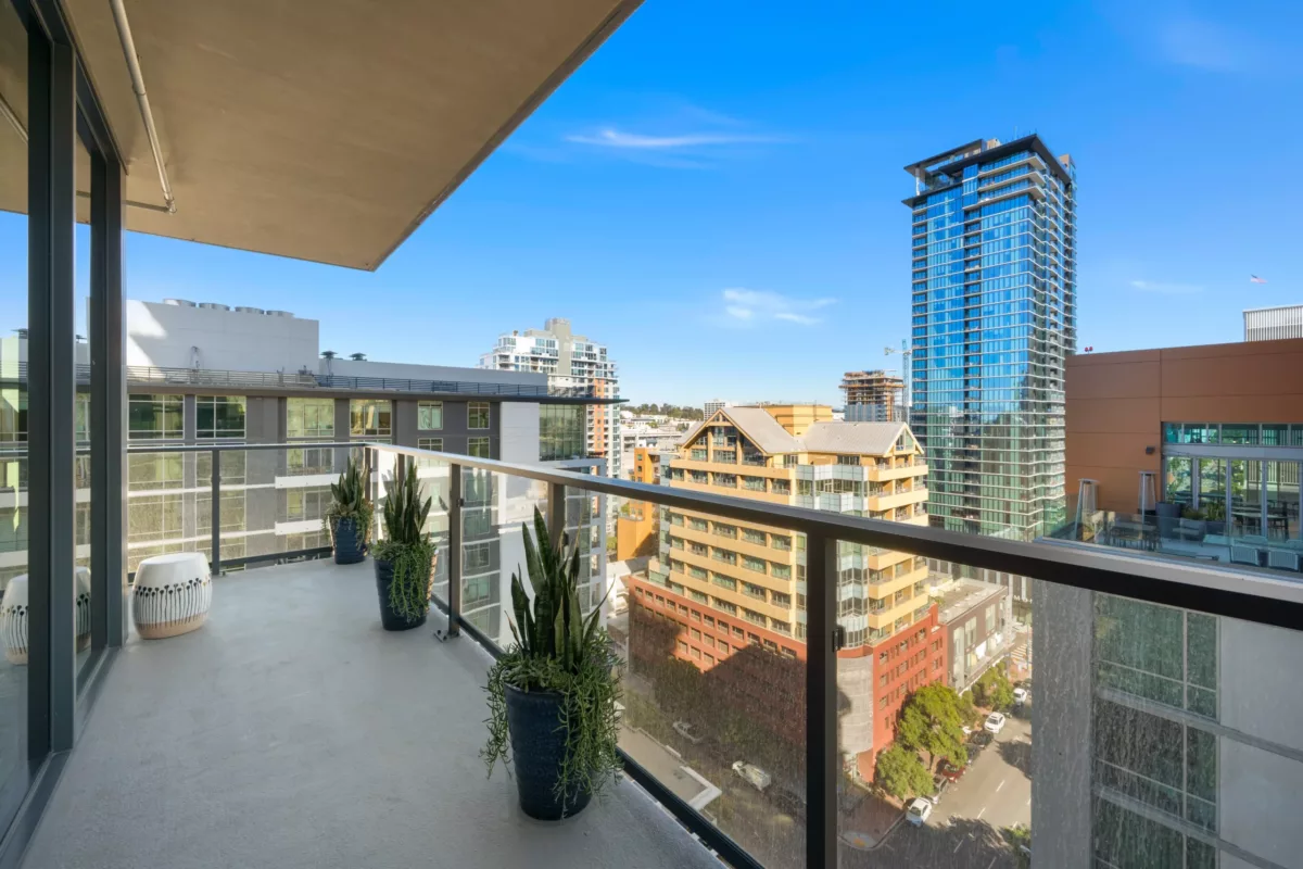 Balcony with potted plants and glass railings, overlooking city buildings under a clear blue sky.