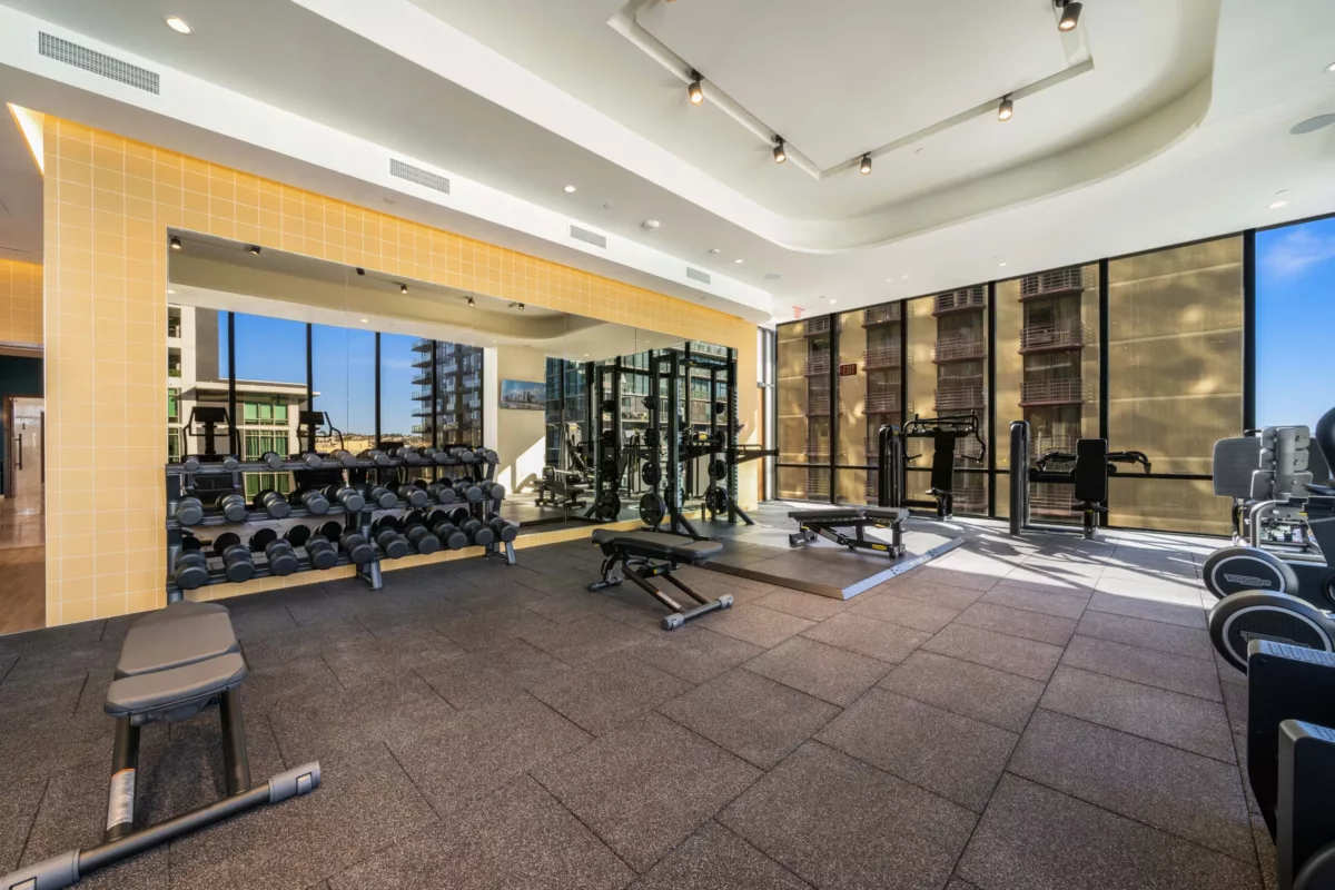Well-lit gym featuring free weights, benches, and a mirrored wall with city views.
