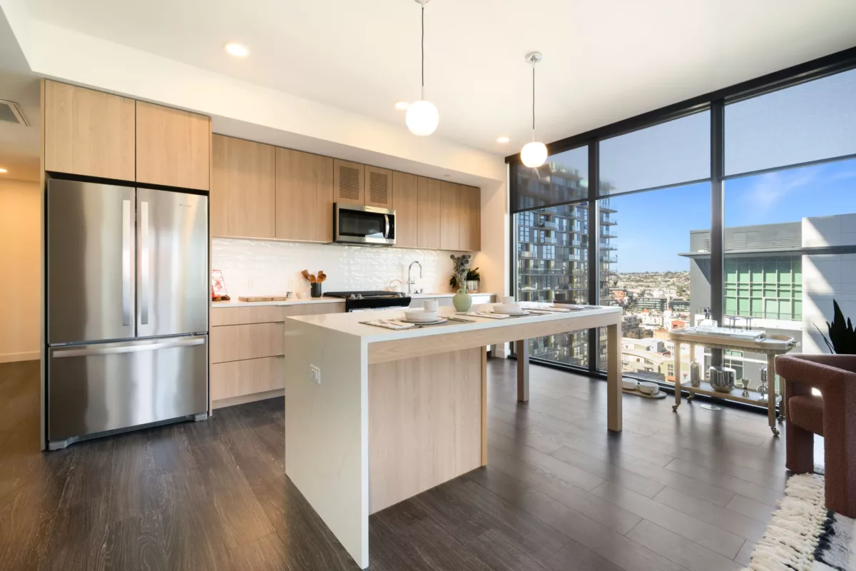 Modern kitchen with light wood-style cabinets, white island, stainless steel appliances, and city views through large windows.