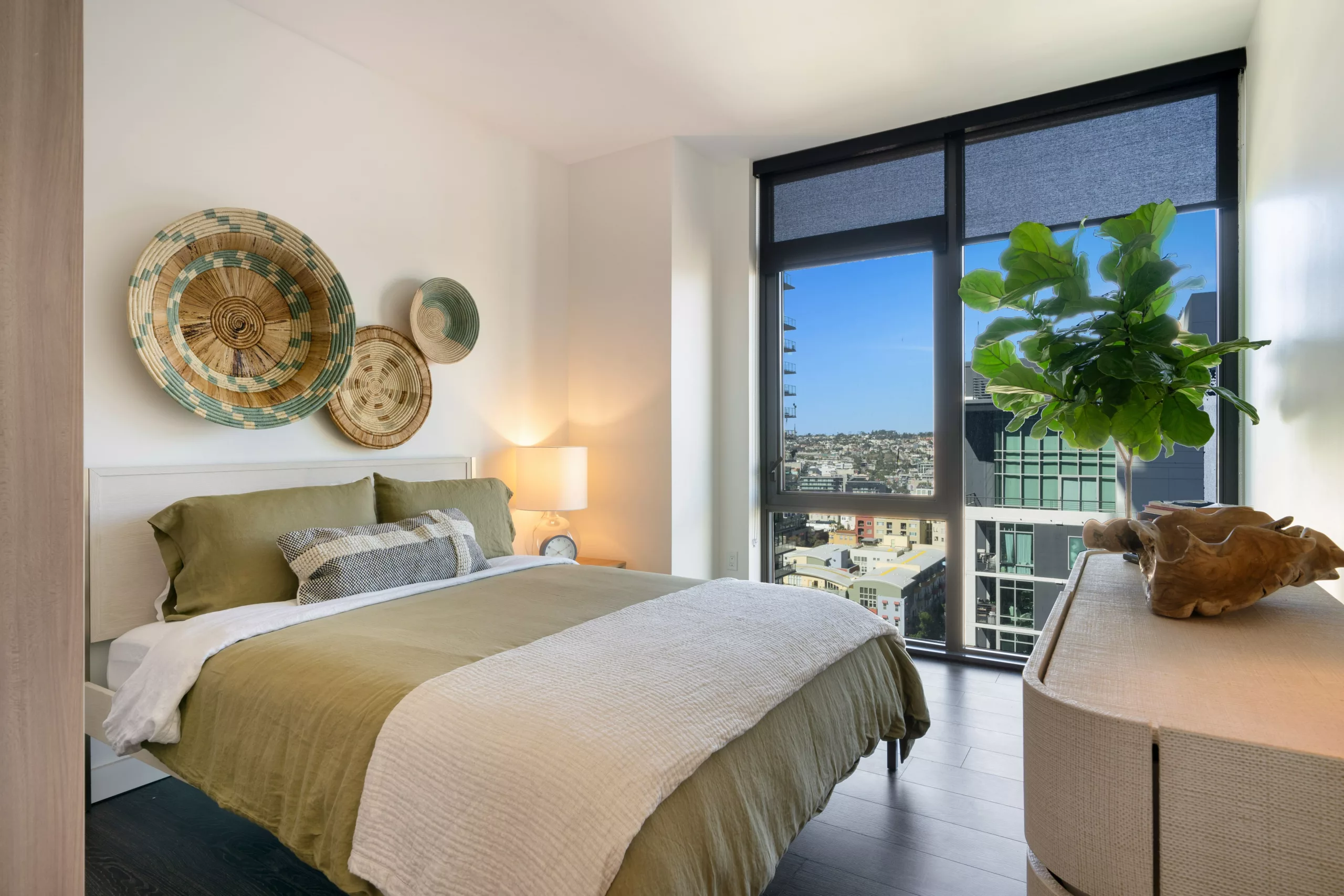 Bright bedroom with green bedding, basket-style wall art, and floor-to-ceiling windows showcasing city views.