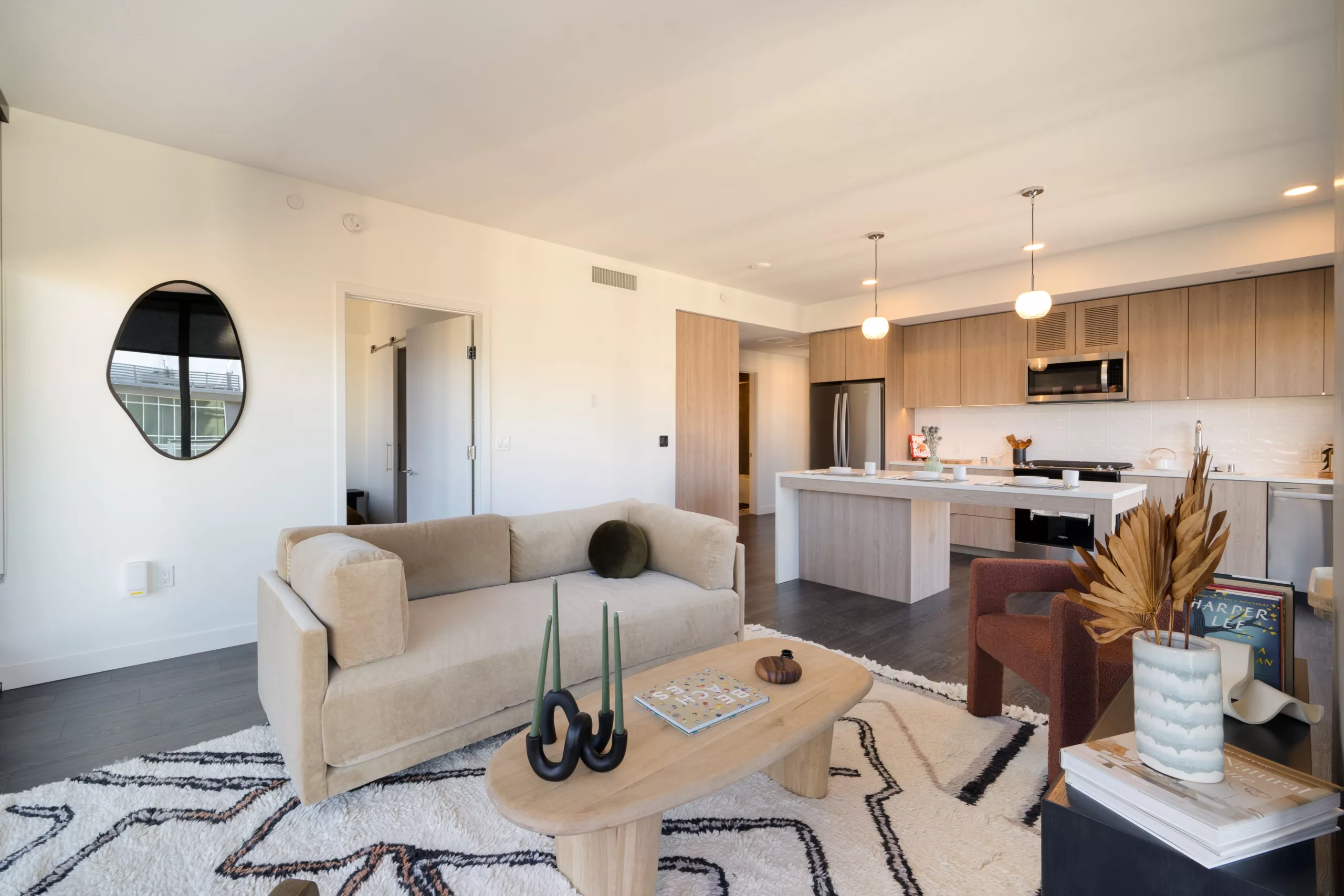 Open-concept living room with a beige sofa, decorative rug, minimalist décor, and light wood kitchen in the background.