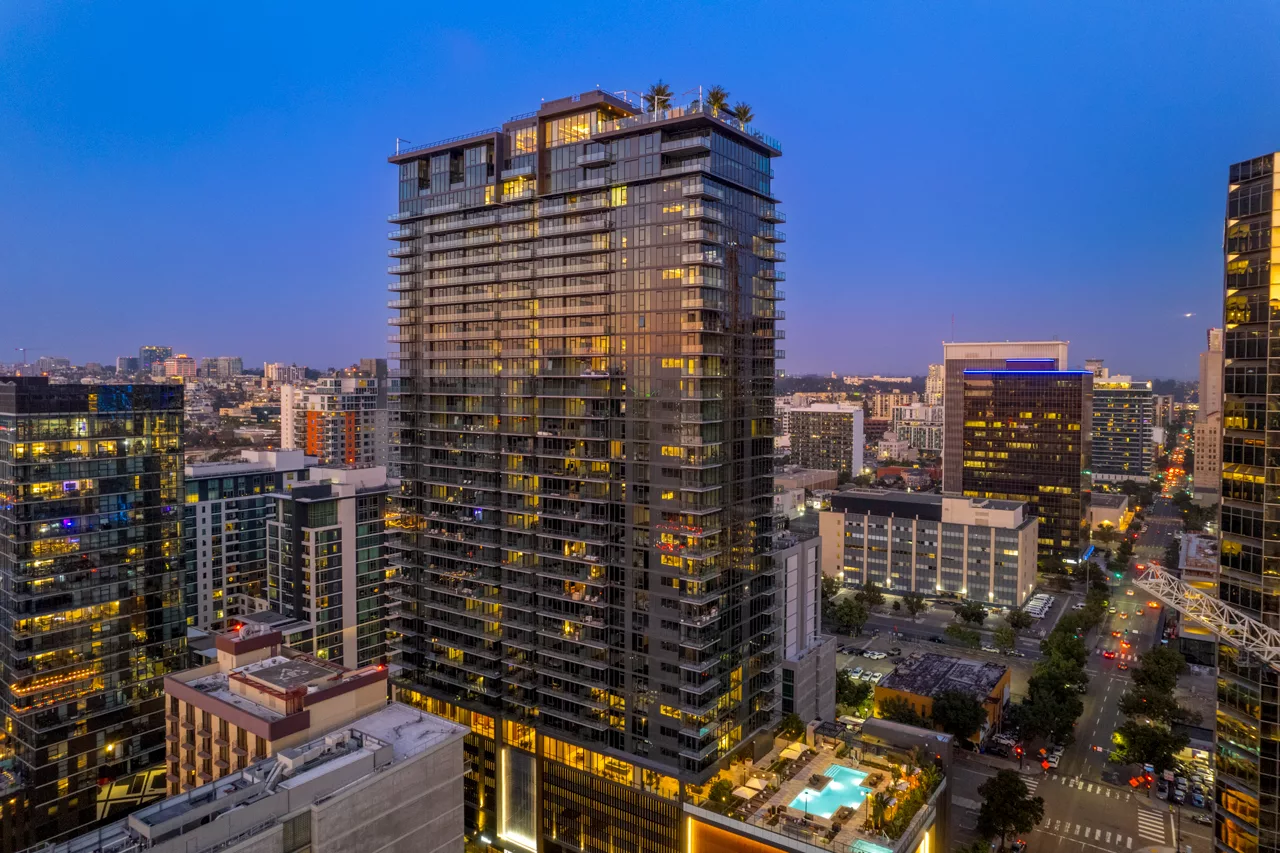 Modern glass high-rise with rooftop trees and pool, lit against a twilight cityscape.